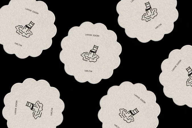 Stylized coasters featuring chair illustrations in water with 'Lagoon Dining' and 'Carlton' text, arranged on a dark background.