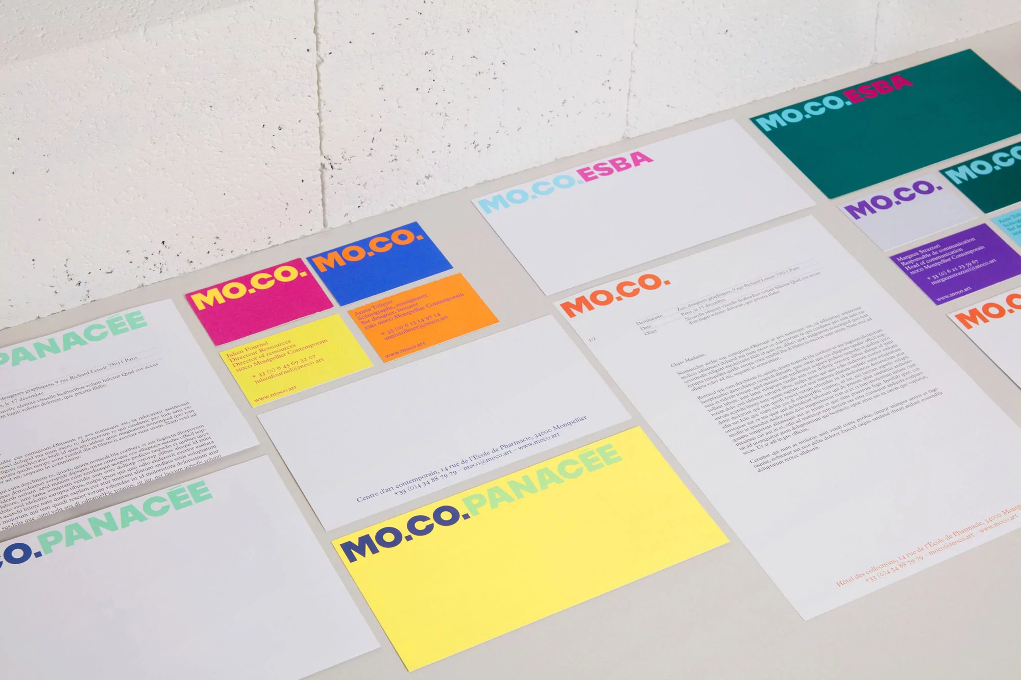 Close-up on colorful letterheads and business cards with the logo MO.CO. PANACEE and MO.CO. ESBA against a neutral white background.