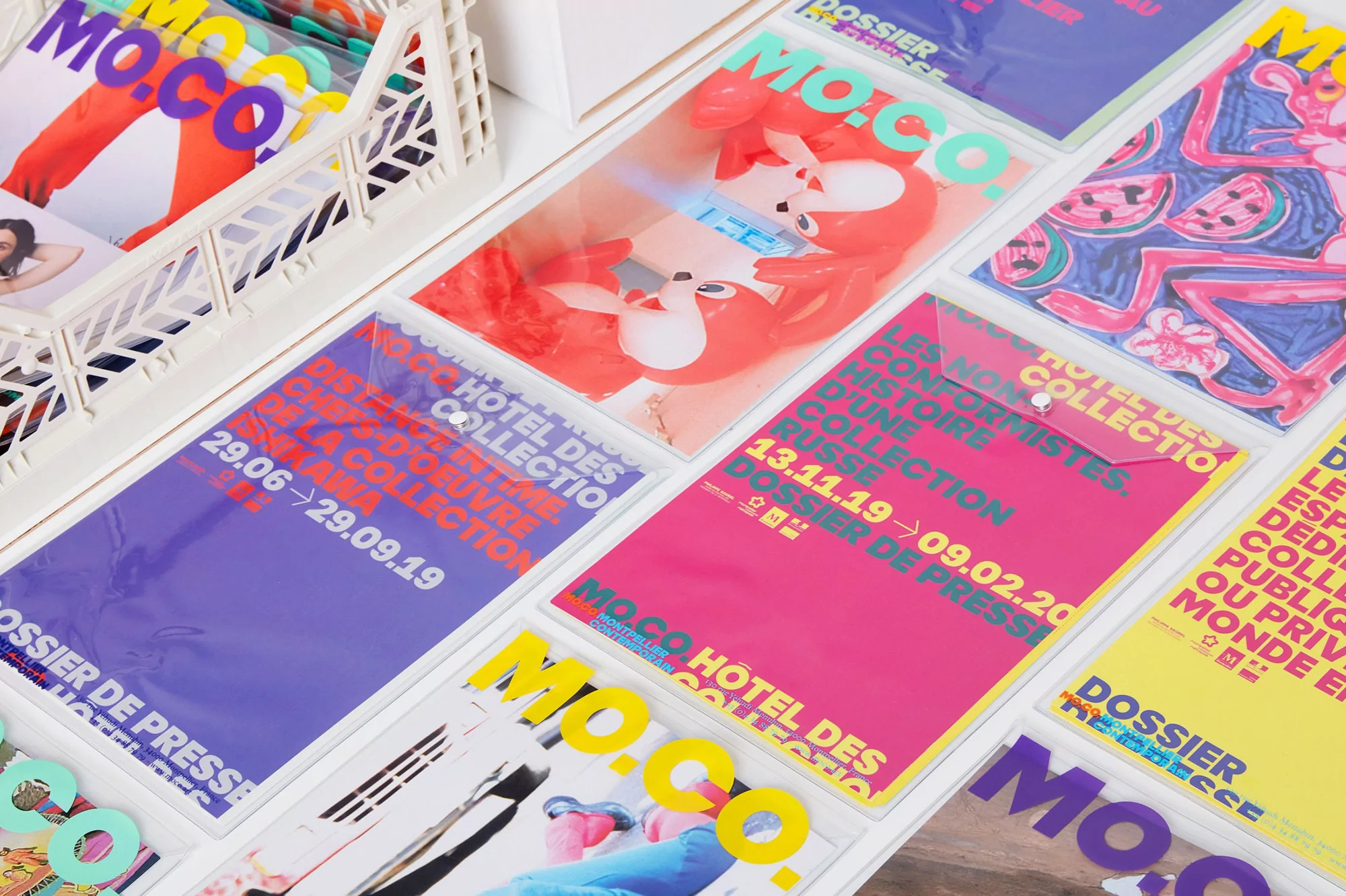 Arrangement of MO.CO. Hotel des Collection press kits with colorful designs in a white basket and spread across a white surface.