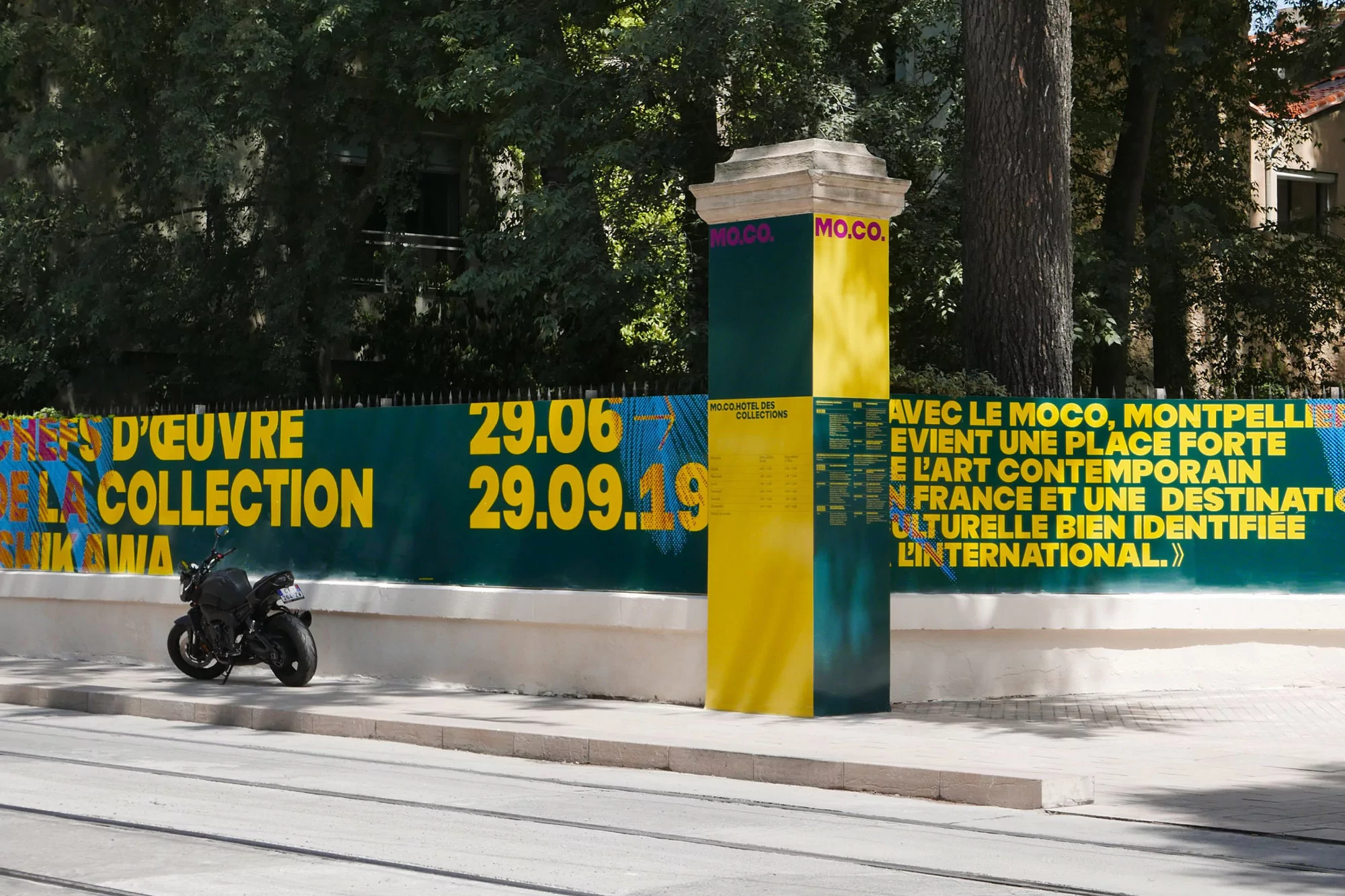 Motorcycle parked near MO.CO. Hotel des Collections exhibition sign in Montpellier, France, with dates and art promotion.