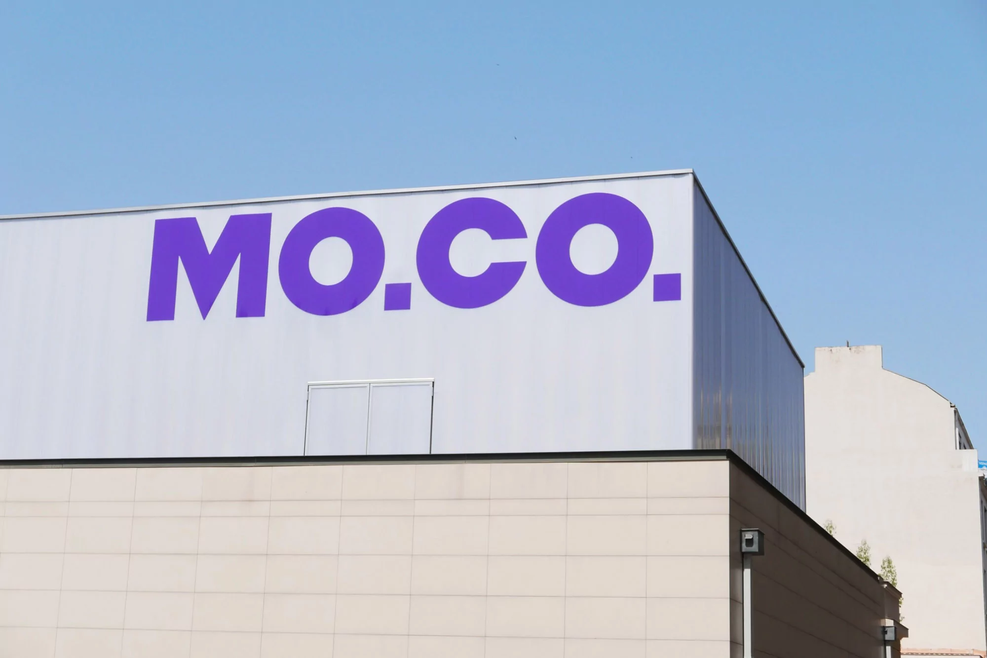 Building exterior featuring bold purple MO CO typography on a white wall against a clear blue sky