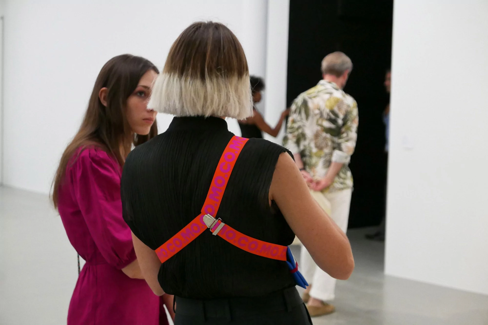 Two women view art at an exhibit, with a blonde bob, black top, and colorful strap harness, set against a white-walled gallery.