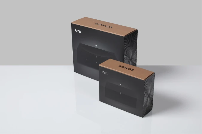 Two Sonos boxes, an Amp and a Port, sit on a white surface against a gray backdrop, showcasing minimalist design and home audio technology.