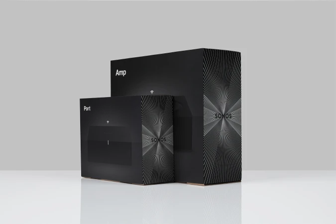 Two black Sonos boxes, one labeled 'Port' and the other 'Amp,' stand on a reflective white surface against a gray backdrop.