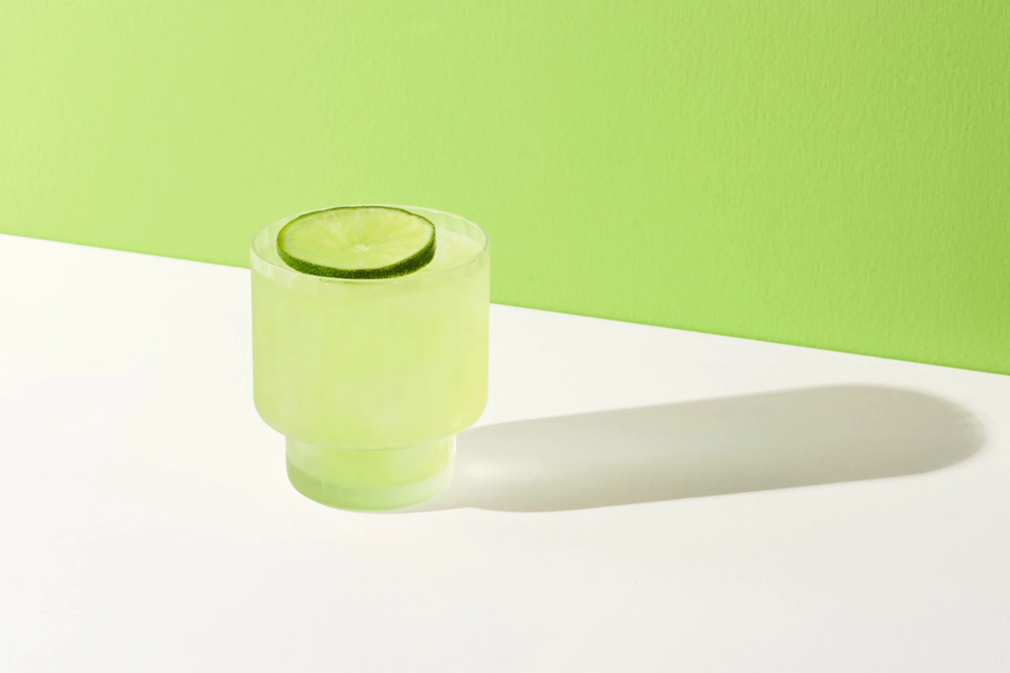 Refreshing light green cocktail in clear glass with lime slice garnish, set against a two-tone green and white backdrop.