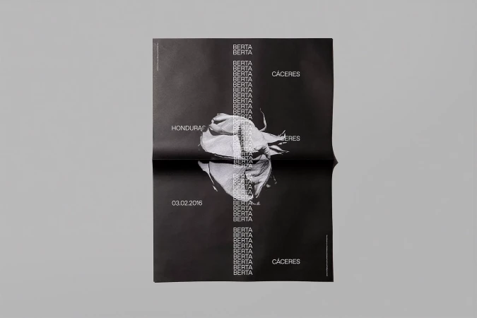 Black poster features a white flower, text repetitions, and Honduras/Cáceres locations. The poster is photographed on a gray surface.