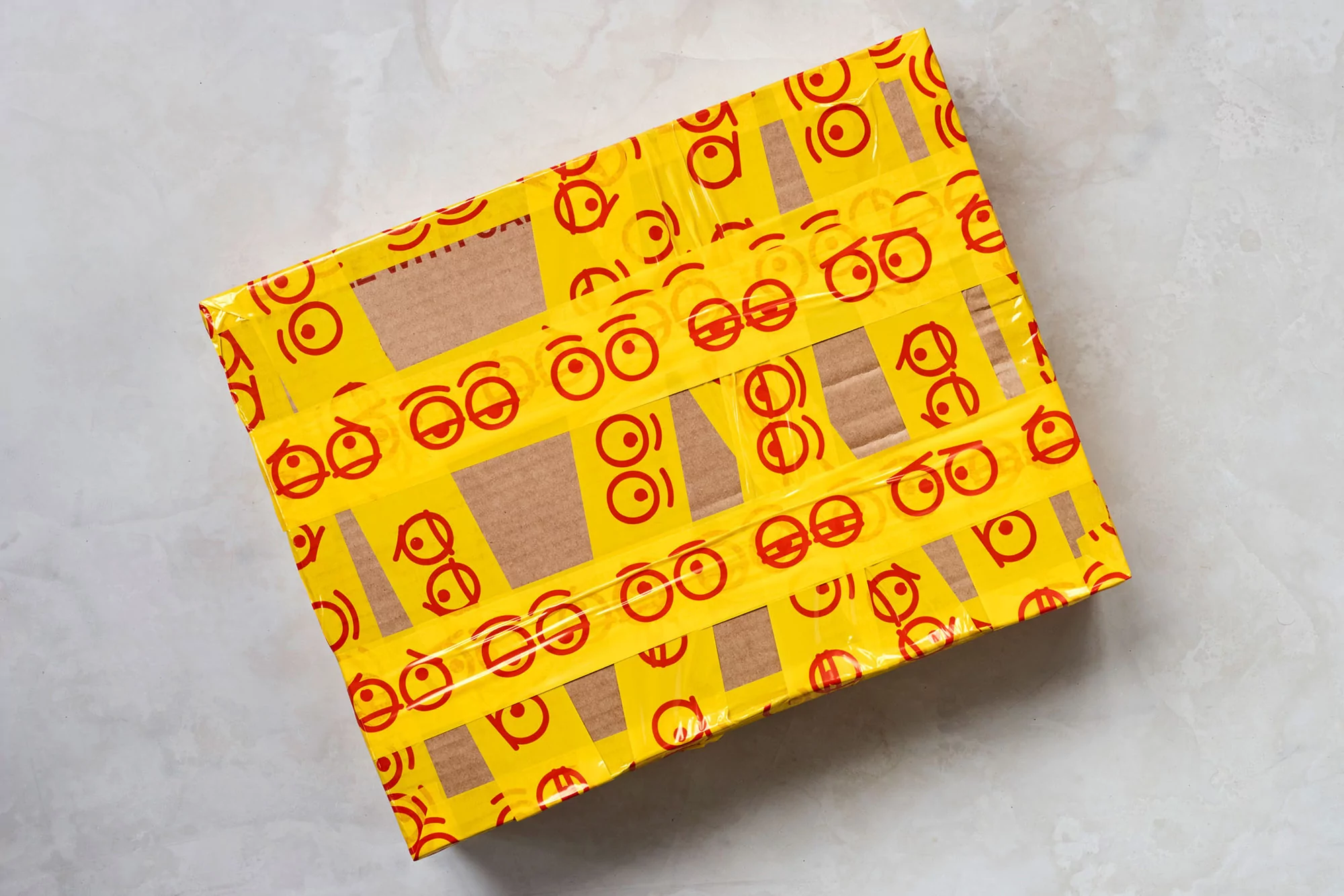 A cardboard shipping box is covered in yellow packing tape featuring red cartoon eye patterns, set on a textured gray surface.