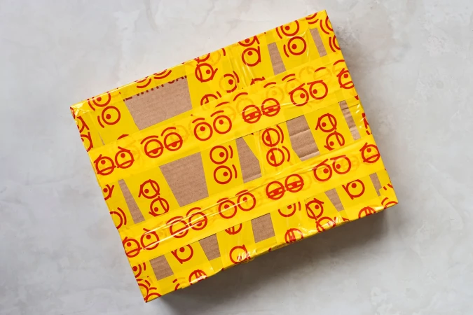 A cardboard shipping box is covered in yellow packing tape featuring red cartoon eye patterns, set on a textured gray surface.