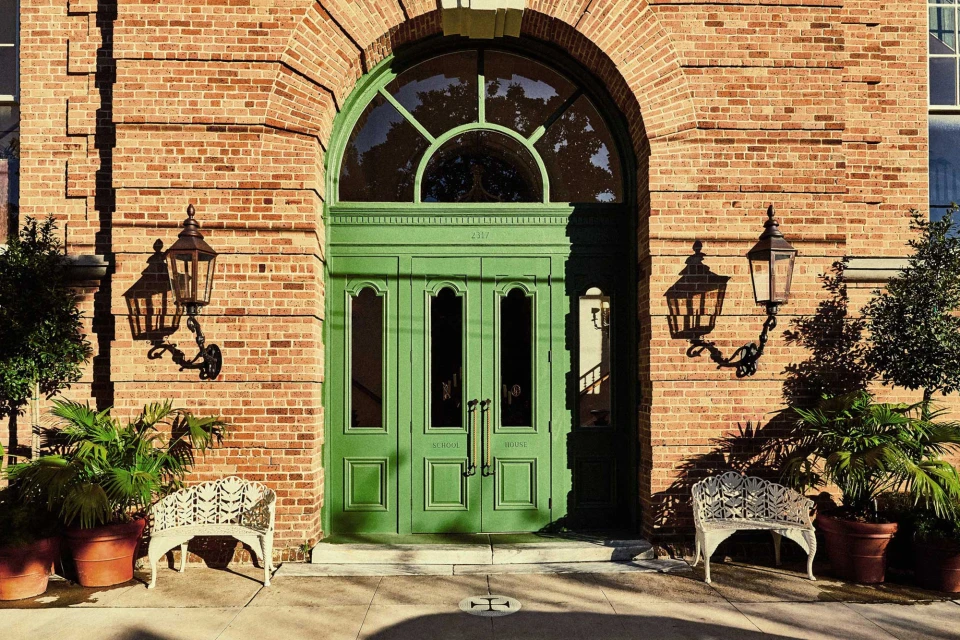 Exterior of a brick building with a green arched doorway flanked by vintage lamps, potted plants, and wrought-iron benches.