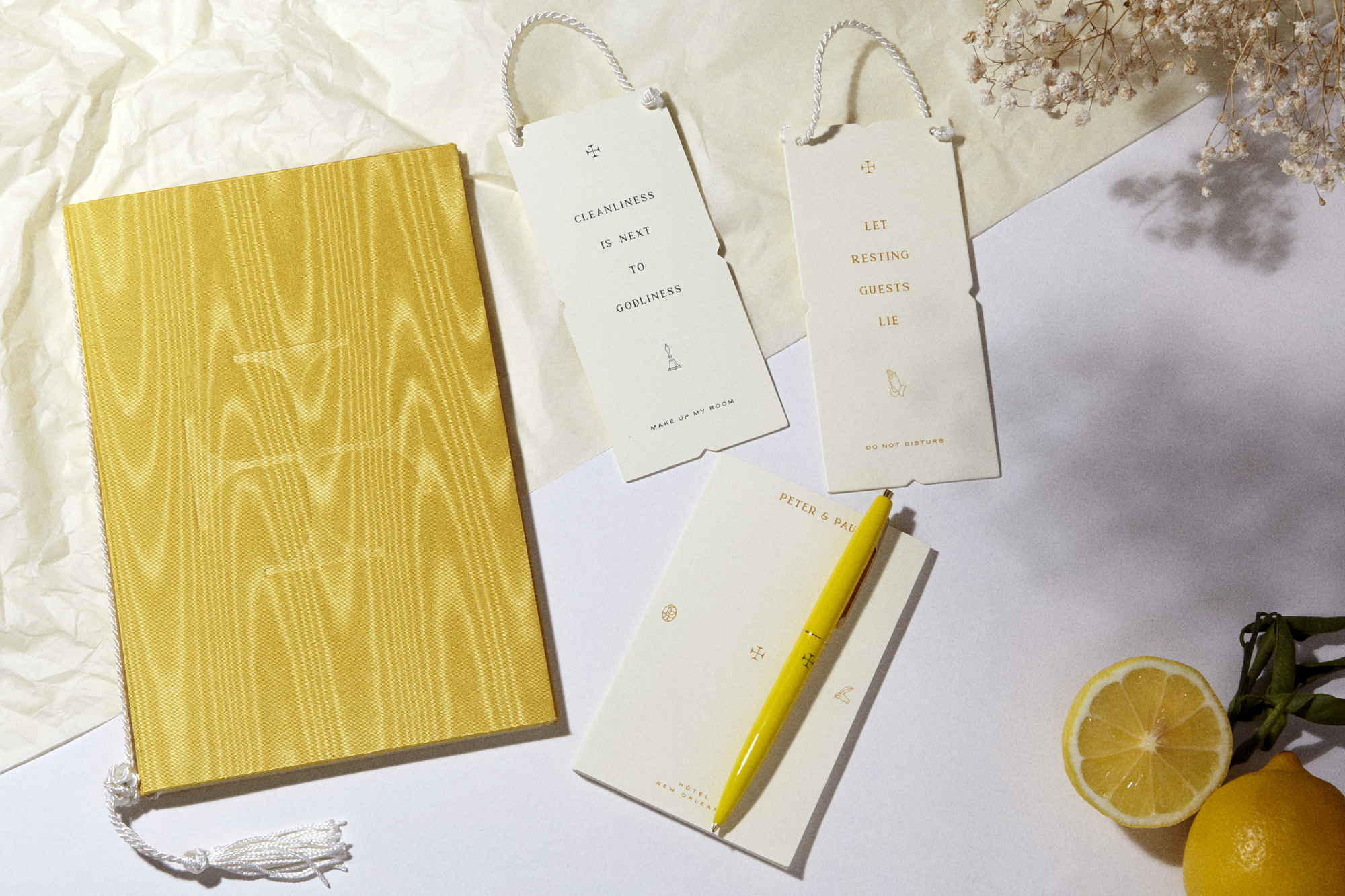 Still life of a yellow journal, pen, notepad, luggage tags, lemons, and baby's breath flowers on a white surface with soft lighting.
