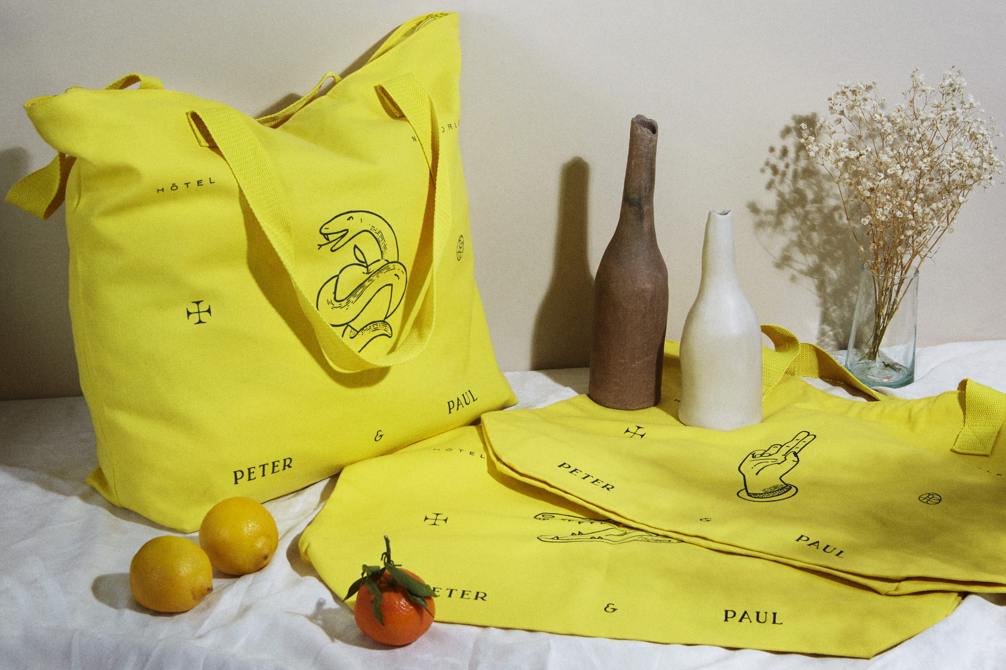 Two yellow tote bags with graphics of a snake and a hand, lemons, orange, vases, and flowers on a white tablecloth.