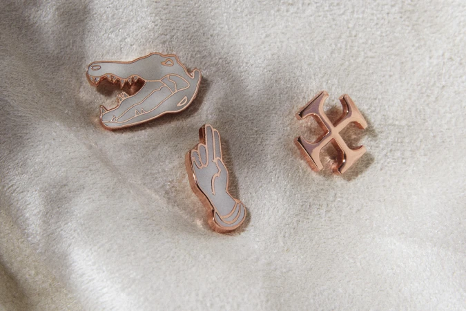 Close-up of three unique enamel pins: a gator head, a hand gesture, and a geometric cross on a soft, textured, off-white surface.