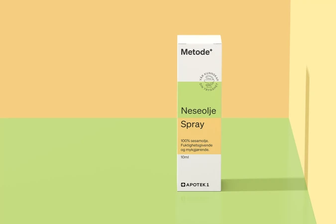 Close-up studio shot of a package of Metode+ Neseolje Spray, a sesame oil product by Apotek 1, against a two-tone yellow and green background.