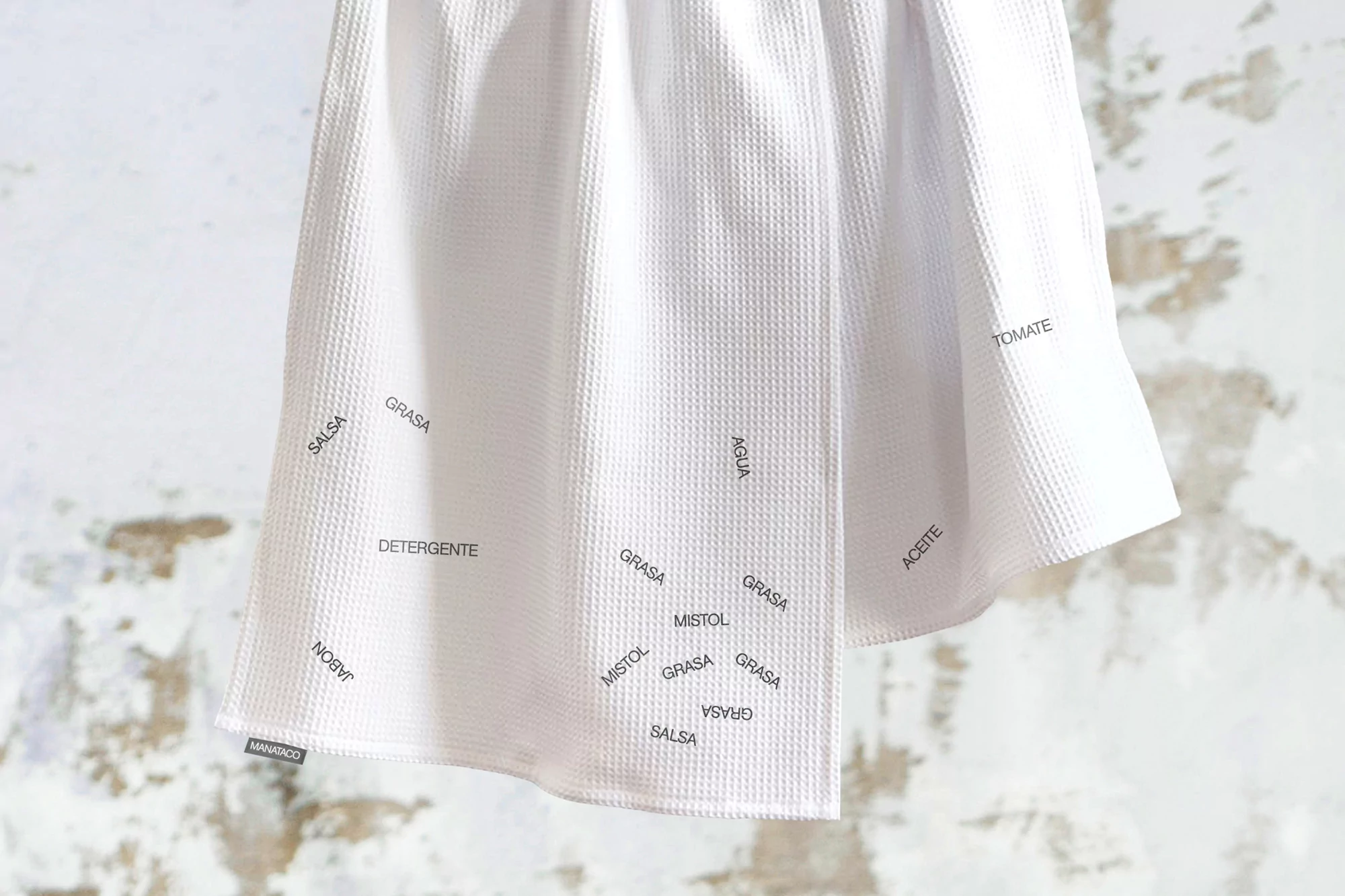 Close-up of a white textured fabric dish towel with printed words in Spanish, hanging against a neutral wall backdrop.