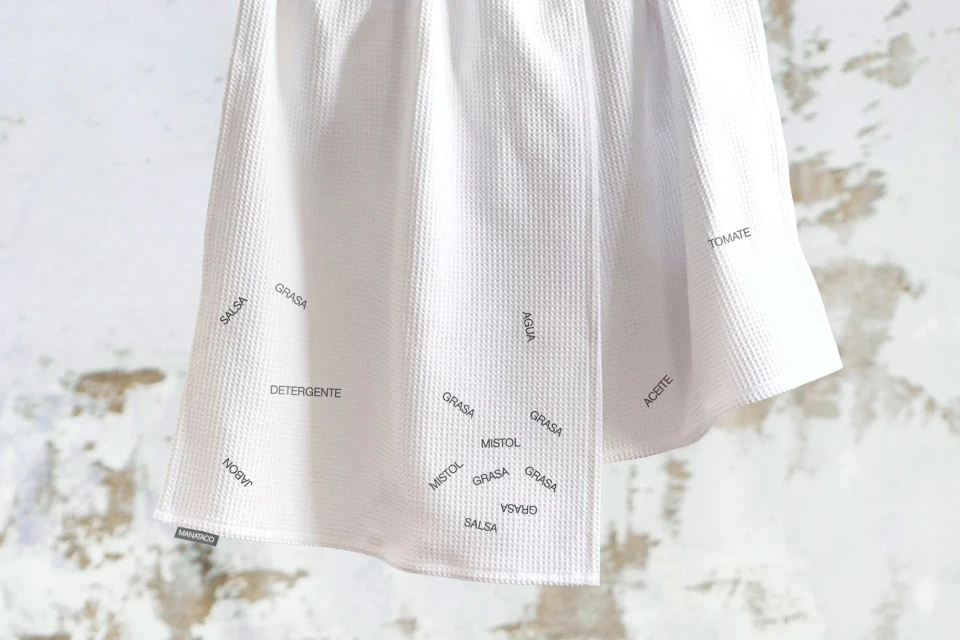 Close-up of a white textured fabric dish towel with printed words in Spanish, hanging against a neutral wall backdrop.