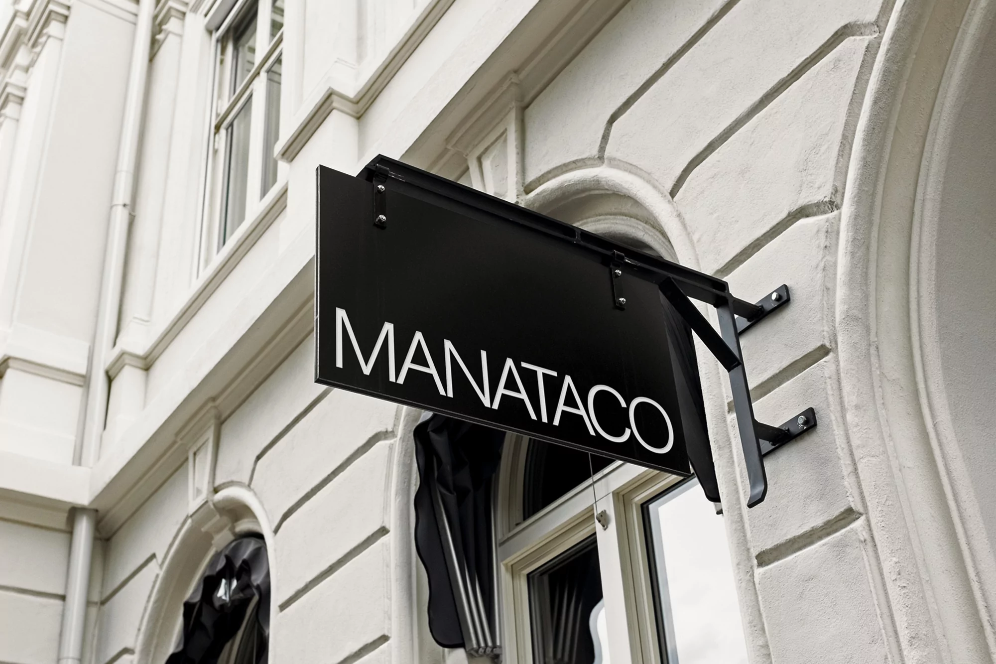 Exterior shot of a black sign with white text 'MANATACO' mounted on a white building with arched windows and architectural details.