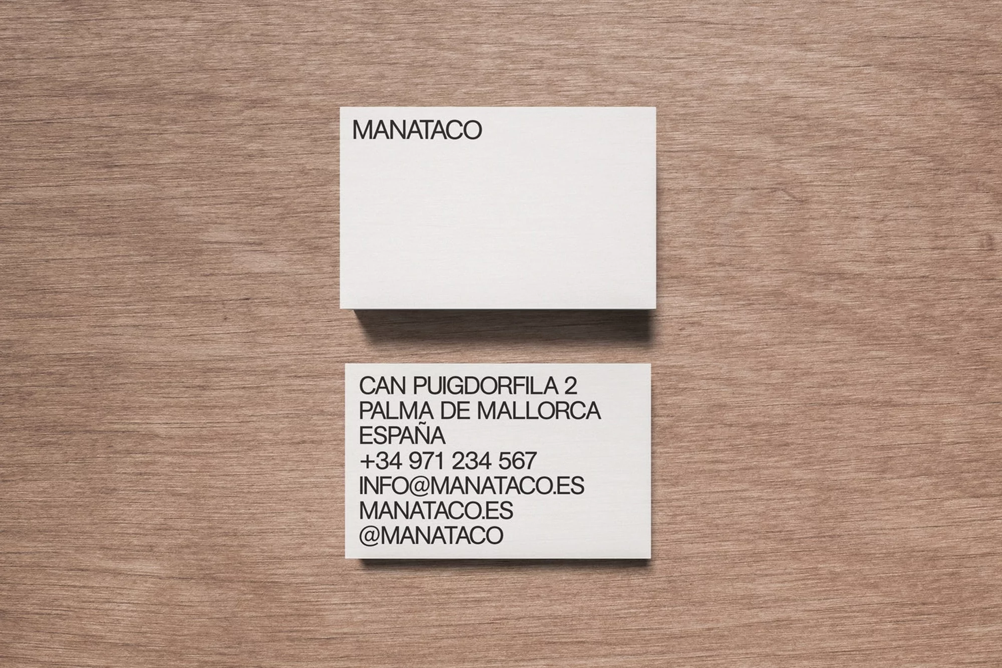 Two white business cards are displayed on a light brown wood table, with contact and company information printed in black ink.