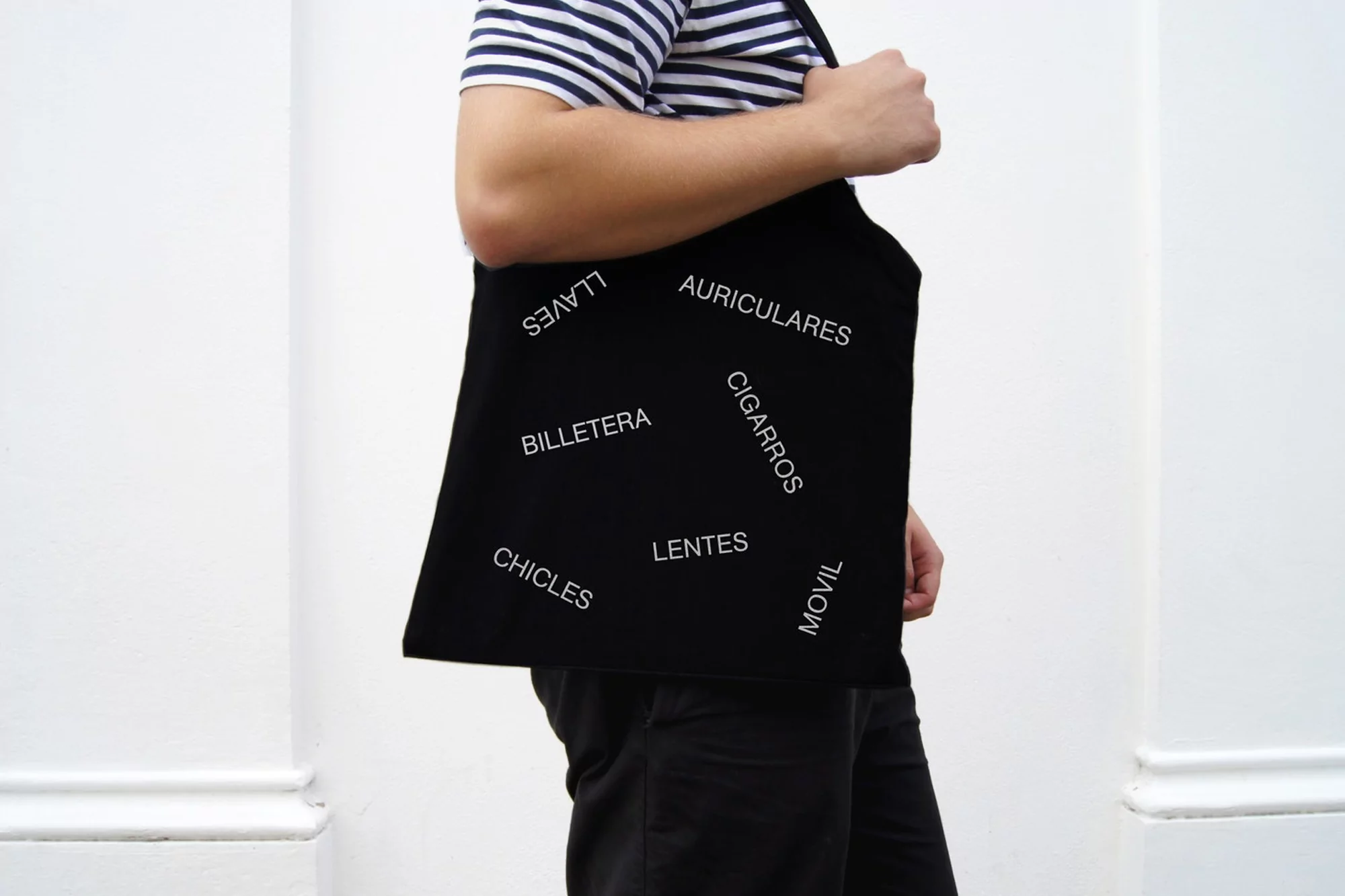 Person wearing a black tote bag with Spanish words printed on it, casual clothing, striped shirt, standing against a white wall.