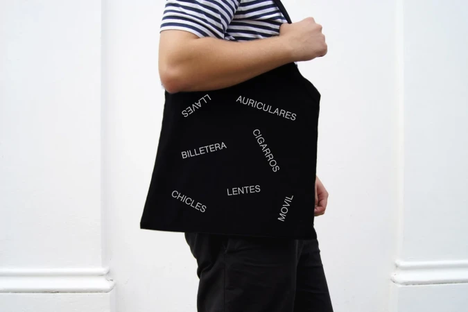 Person wearing a black tote bag with Spanish words printed on it, casual clothing, striped shirt, standing against a white wall.