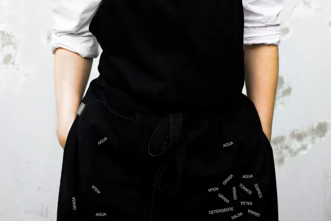 Person wearing a black apron with kitchen-related words printed on it, standing against a textured white wall, in a studio shot.