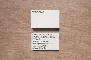 Two white business cards resting on a textured wooden surface, displaying minimalist black typography for Manataco.