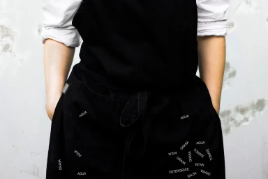 Person wearing a black apron with typography over a white shirt against a textured concrete wall.