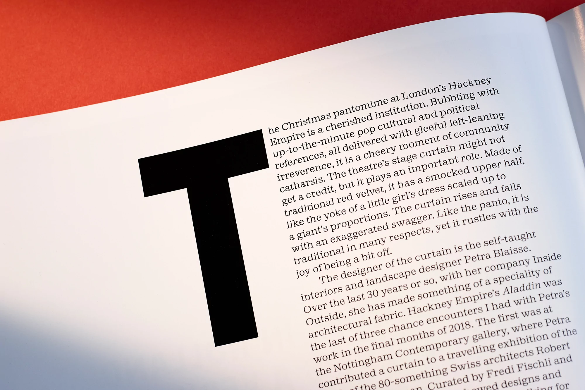 Close-up of an open book with a black sans-serif letter 'T' and surrounding text on a white page, against a red background.
