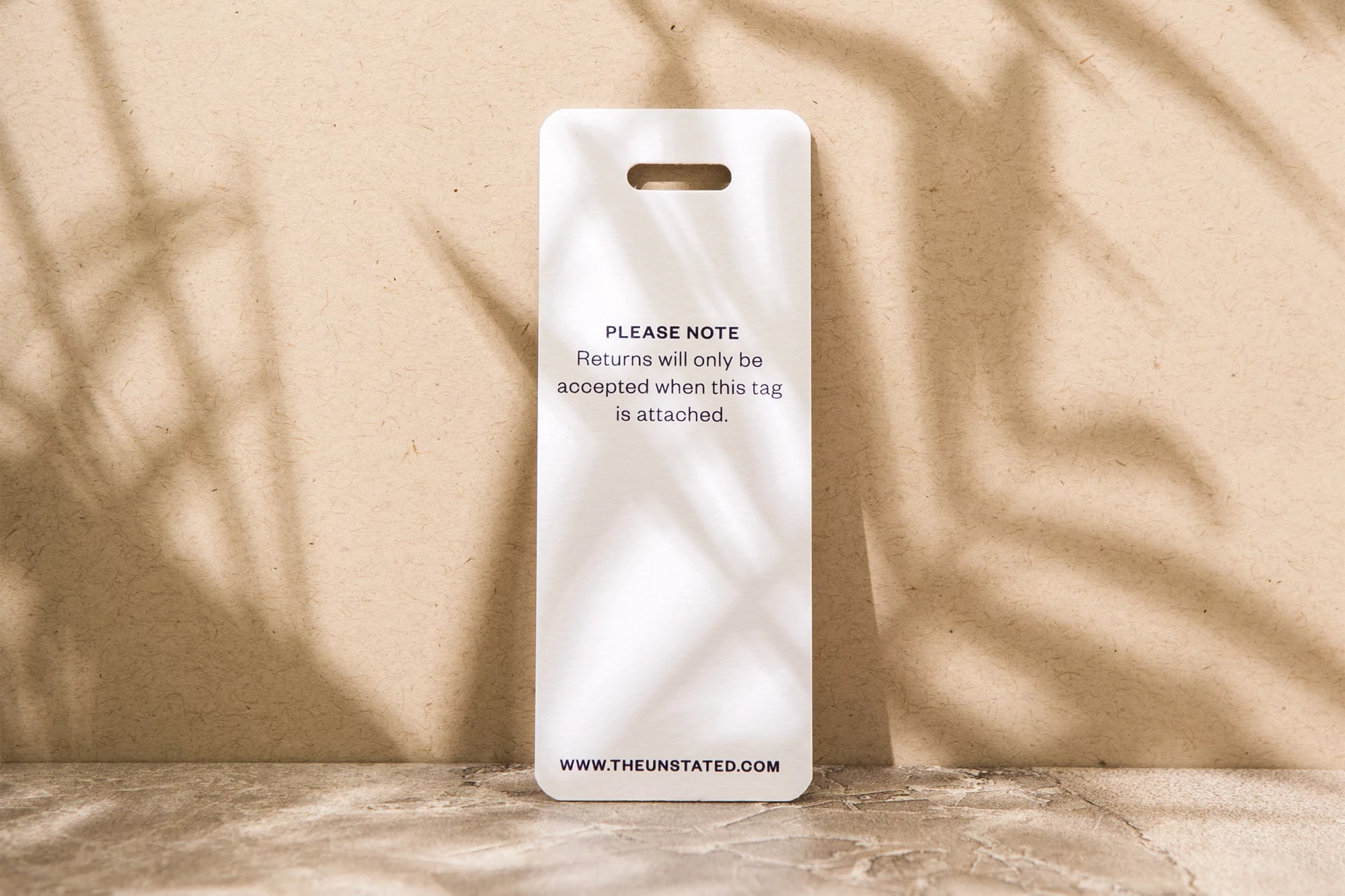 White tag with rounded corners and a slot at the top with a brand's return policy statement, sitting on a neutral surface.
