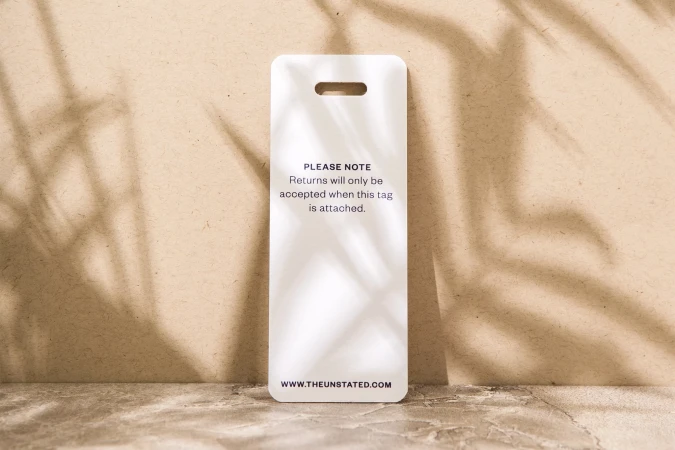 White tag with rounded corners and a slot at the top with a brand's return policy statement, sitting on a neutral surface.