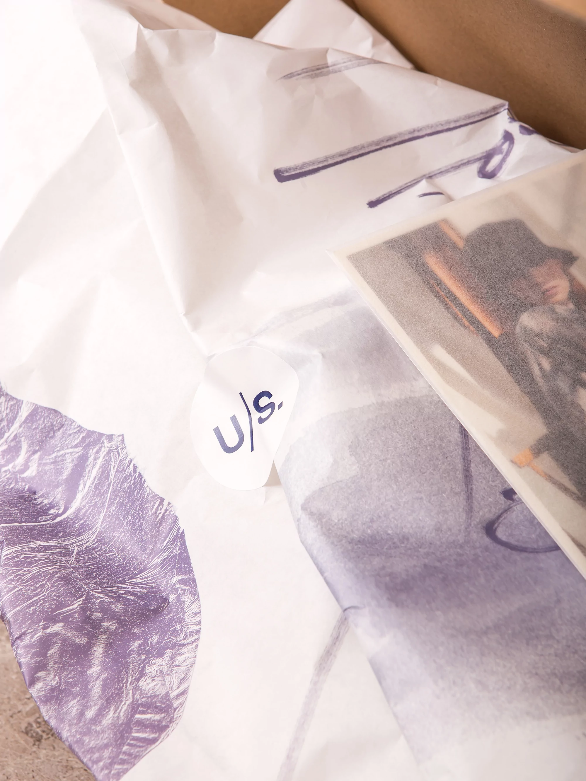 Image of white tissue paper with a blue logo and a color print featuring a woman in a hat, all arranged on a neutral surface.