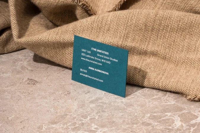 Close-up of a blue business card resting on a stone surface, with a beige, textured fabric draped in the background.