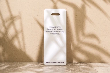 White branded hang tag placed upright against a textured beige surface with soft leaf shadow patterns.