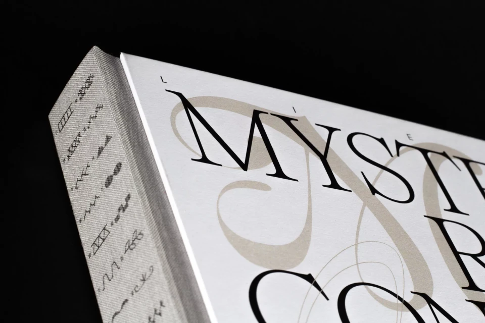 Close-up of book cover with black 'Mystery' title on white and gray cover; gray, geometric pattern on book spine; black background.
