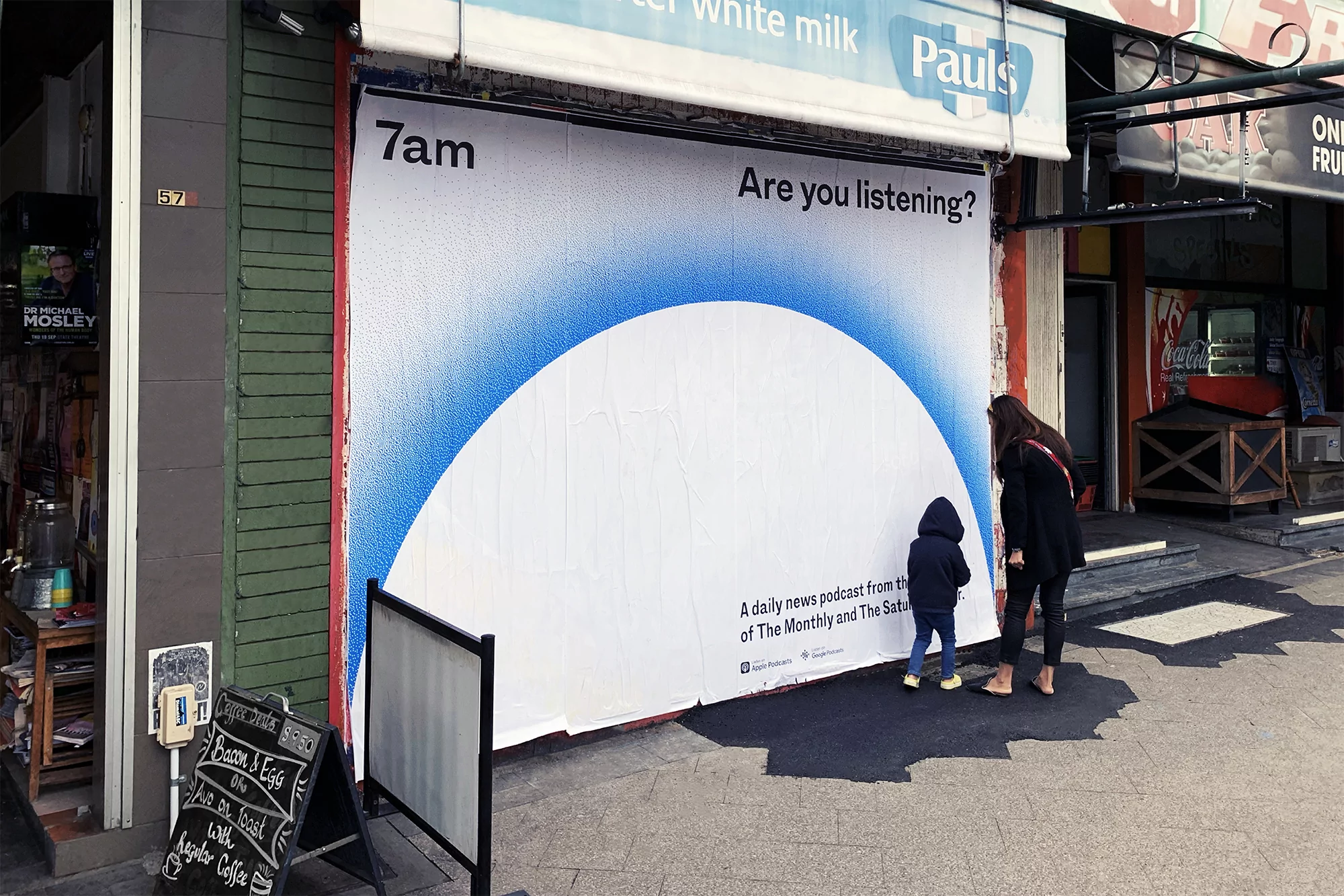 Outdoor ad featuring a podcast promotion, with a woman and child looking at the ad on a building exterior in an urban setting.