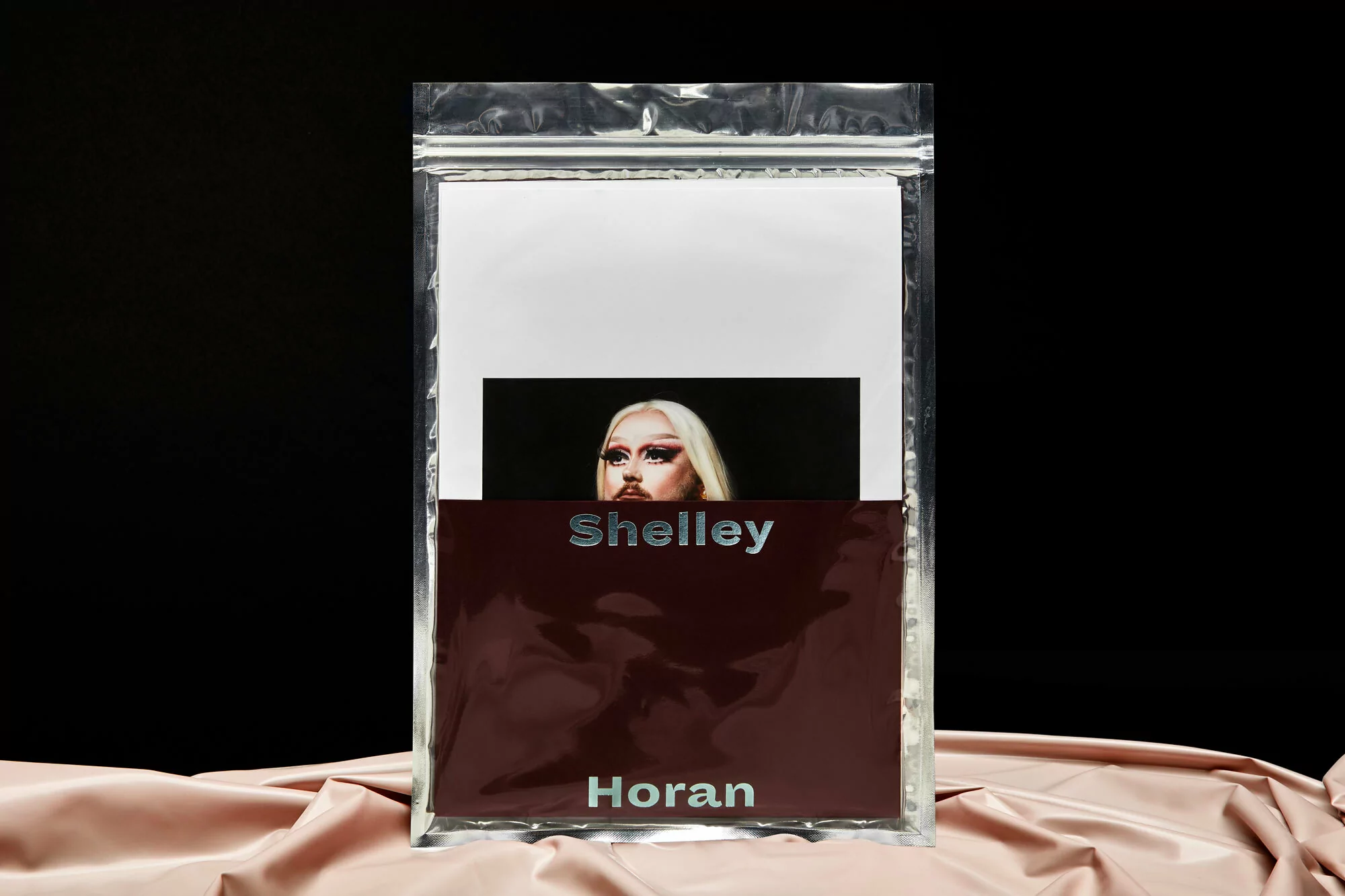 Shelley Horan image inside a plastic sleeve on a light pink fabric. The image features a person with blond hair and makeup.