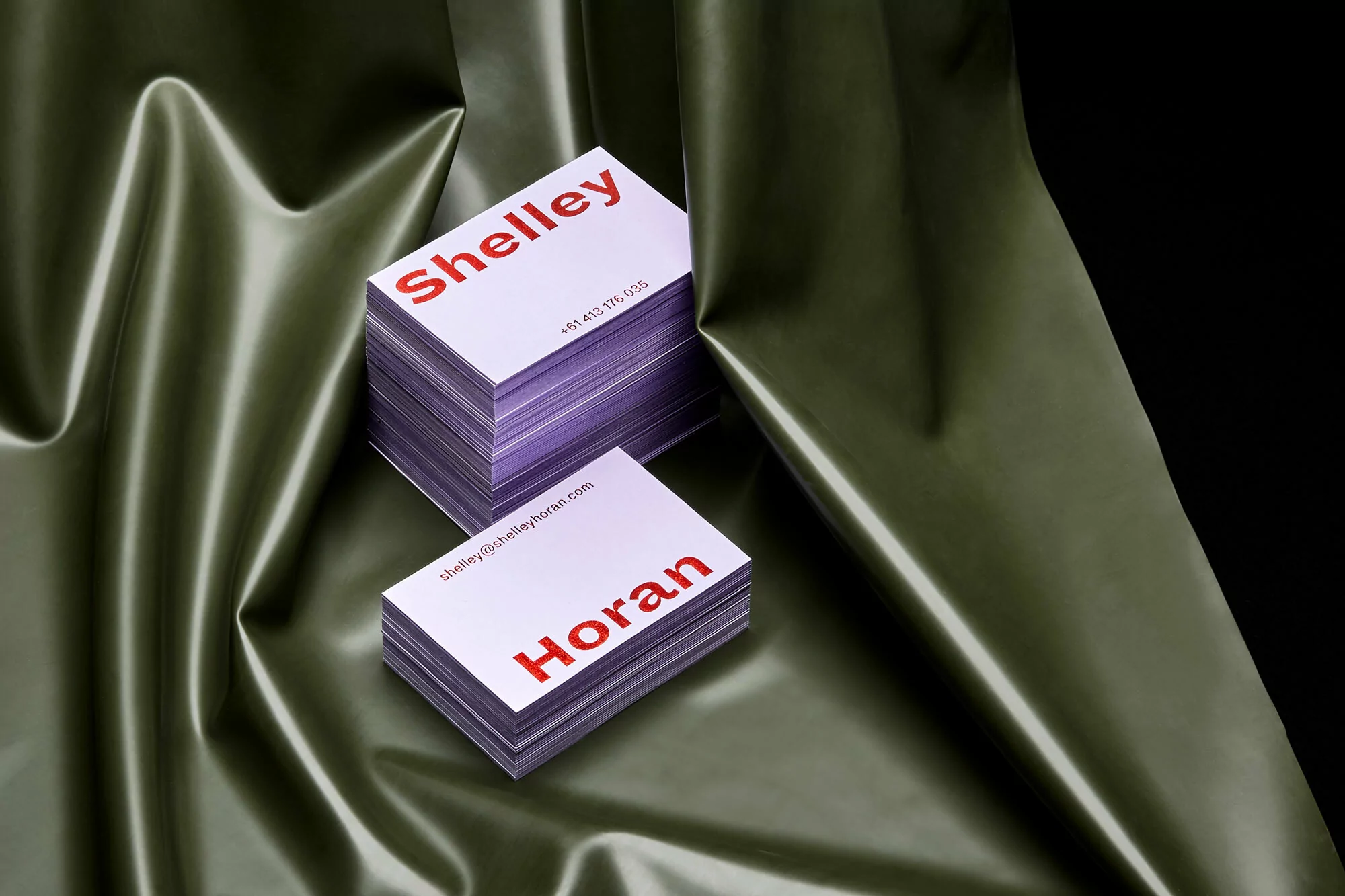 Stacks of business cards with names and contact details against a draped olive-green satin backdrop, creating an elegant professional presentation.