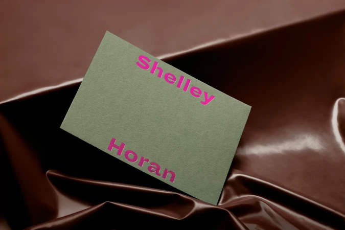 Olive-green card with the words 'Shelley Horan' in a bright pink font, displayed on a textured brown satin background. 