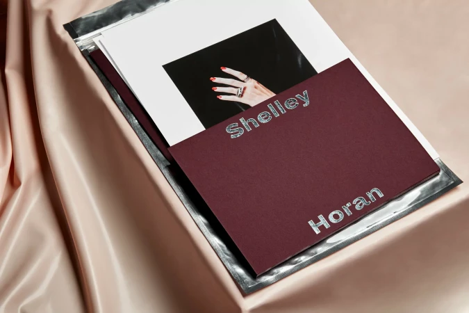 Close-up still life: photograph of a hand with rings is inside a metallic envelope with a dark red card bearing the names Shelley Horan.