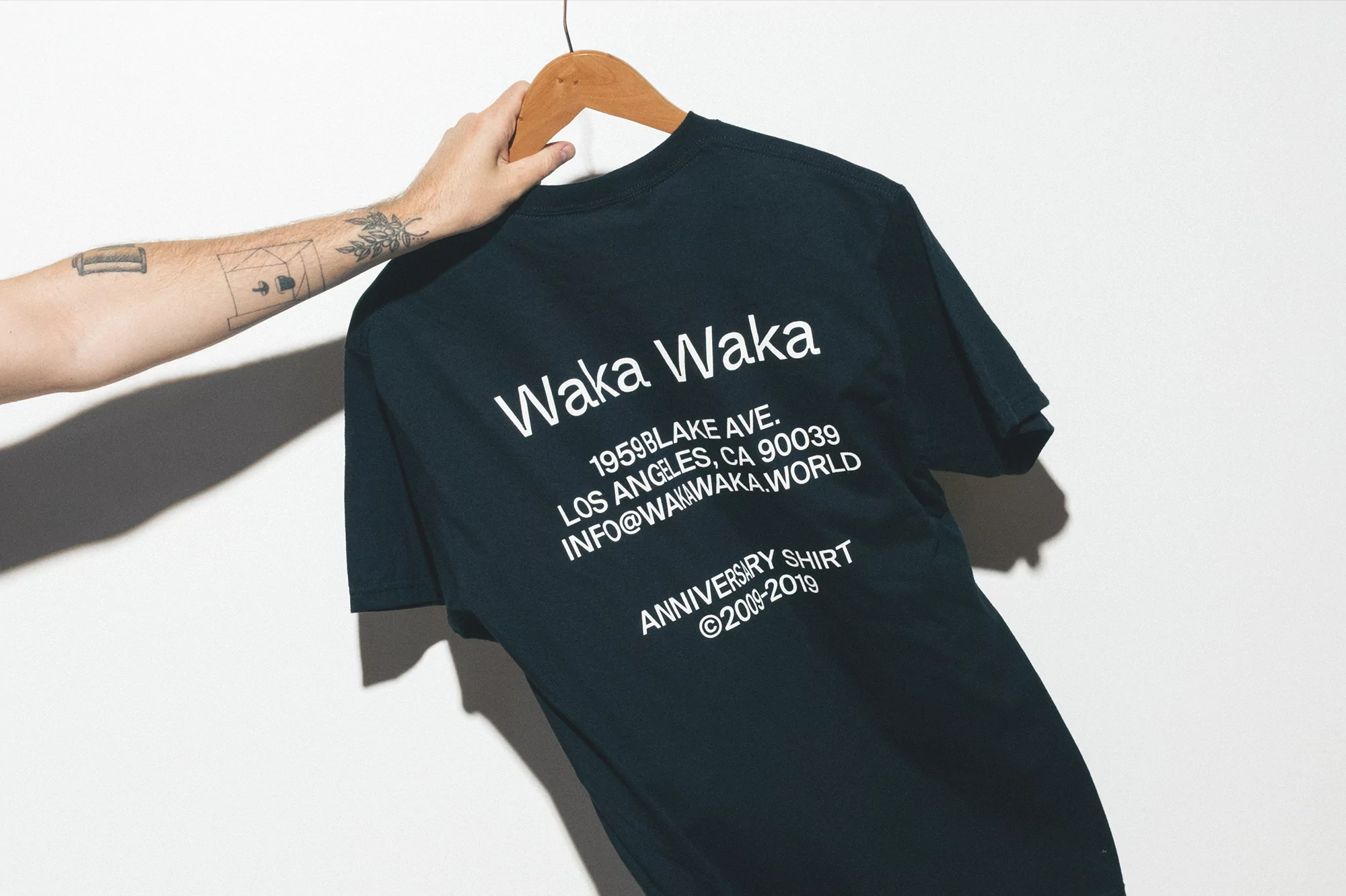 Dark-colored T-shirt on a wooden hanger, held by a hand, displaying brand text and address against a white background in studio lighting.