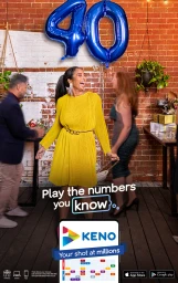 A laughing woman in a yellow dress standing in front of a red brick wall beneath large blue forty balloons