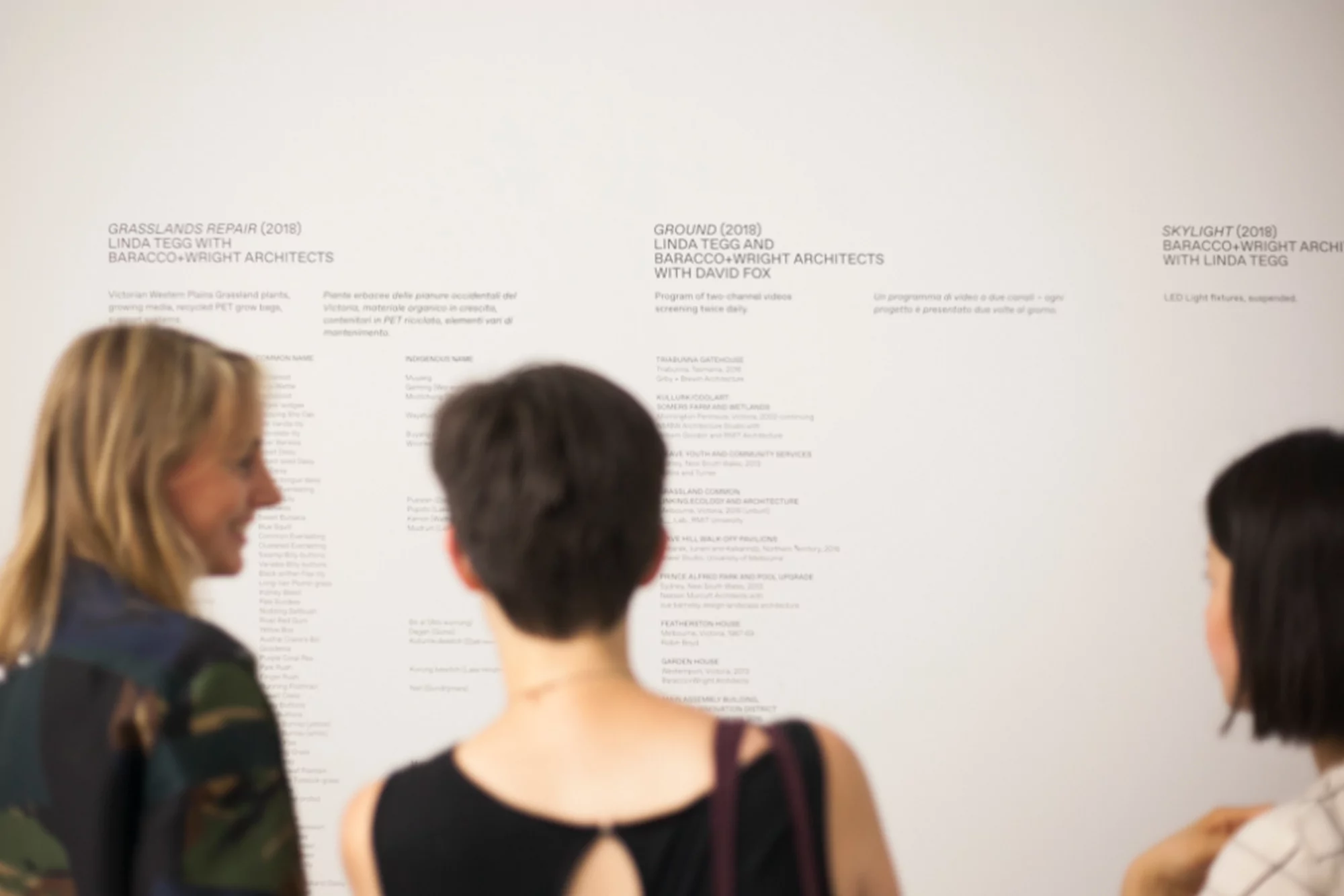 Three women are standing in front of a white wall looking at architectural plans listed. The women are blonde, brunette and have dark hair.