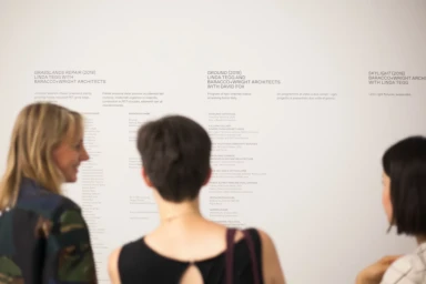 Three visitors viewing exhibition wall text with project titles for the Australian Pavilion Brand Identity.