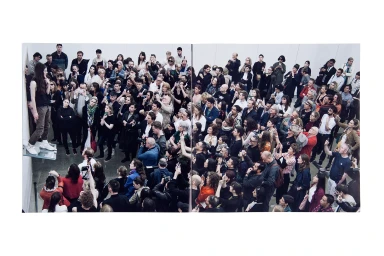 High-angle view of a dense crowd gathered around a gallery space, watching a performer standing elevated against a white wall.