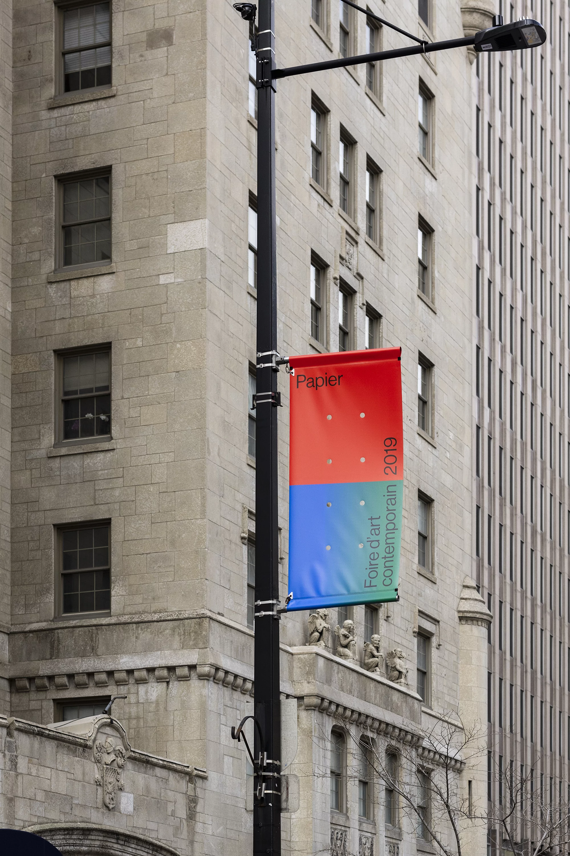 Cityscape featuring a lamp post displaying an art fair banner for Papier 2019, with the backdrop of vintage architecture.