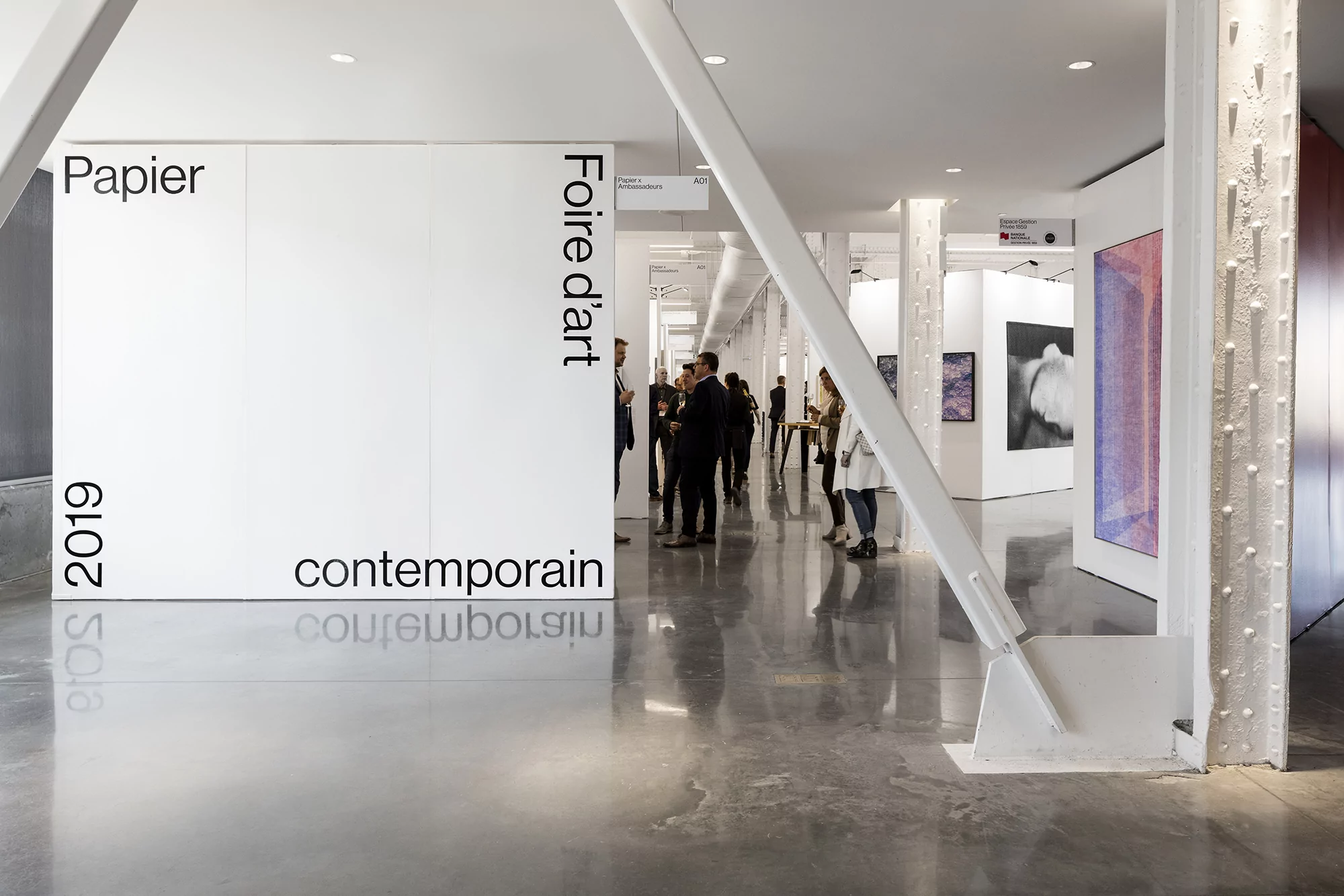 Art fair exhibit featuring crowds of people viewing contemporary art, with a polished concrete floor and white walls and structural elements.