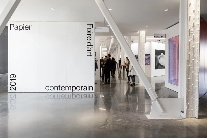 Art fair exhibit featuring crowds of people viewing contemporary art, with a polished concrete floor and white walls and structural elements.