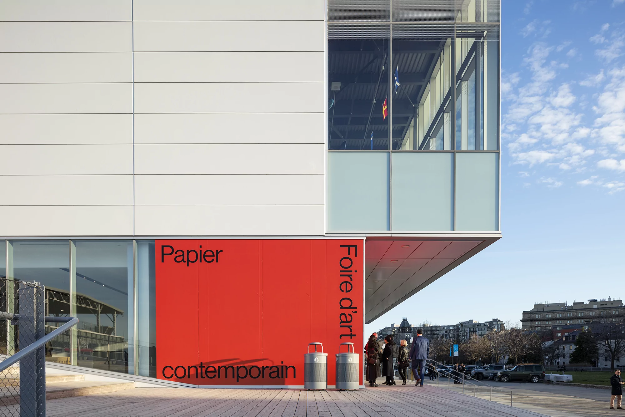 Exterior view of modern building with large windows and bold text signage featuring the 'Papier' art fair, with people outside.