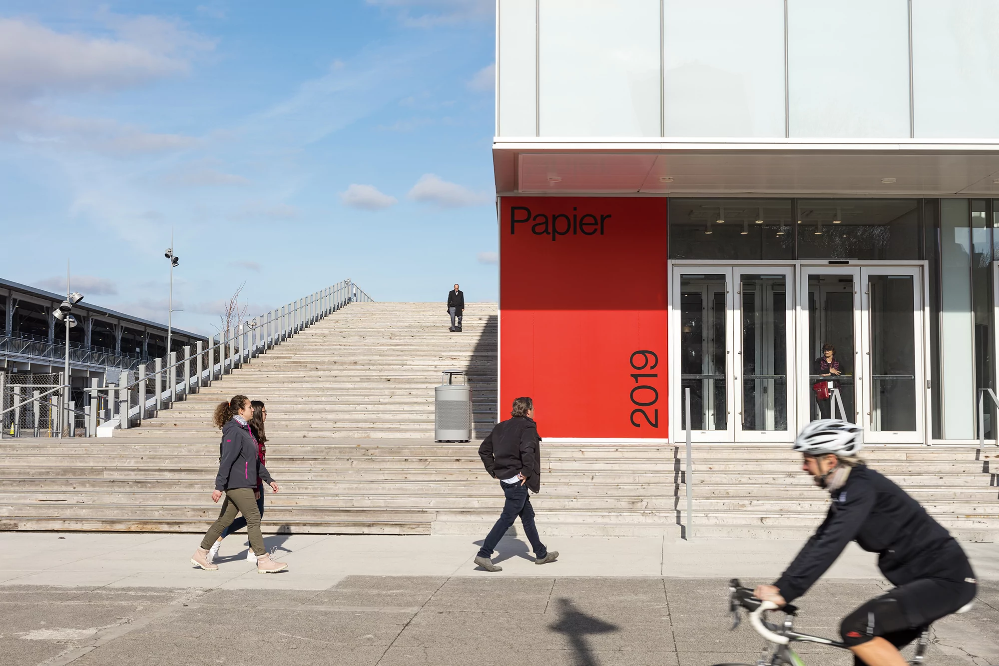 Modern urban scene with people walking and cycling near a building with a large red sign reading 'Papier 2019' on a sunny day.