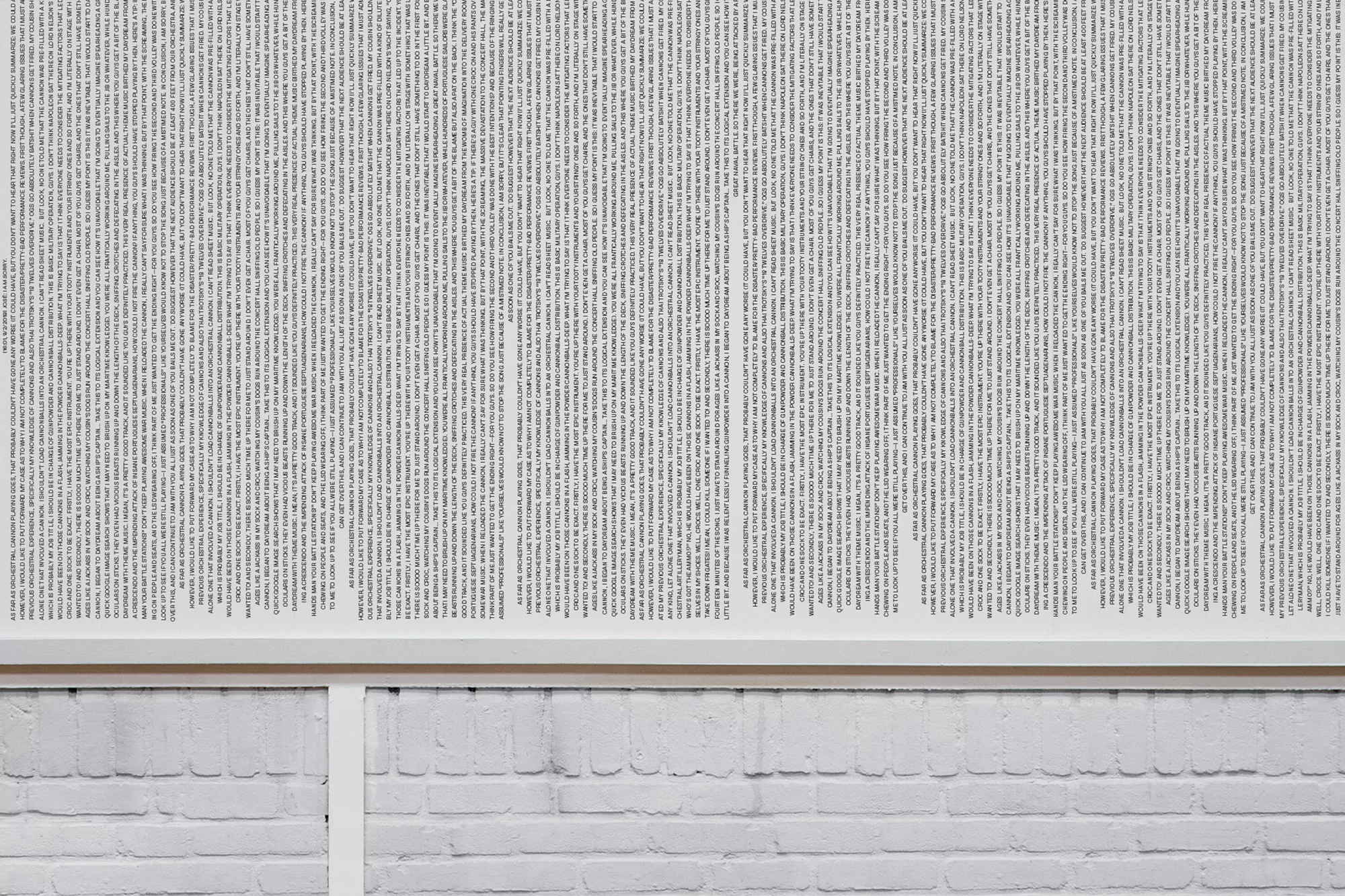Abstract art features dense columns of text, arranged vertically on a white background, mounted above a white painted brick wall.