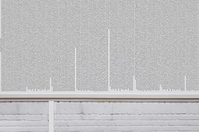 Abstract art features dense columns of text, arranged vertically on a white background, mounted above a white painted brick wall.
